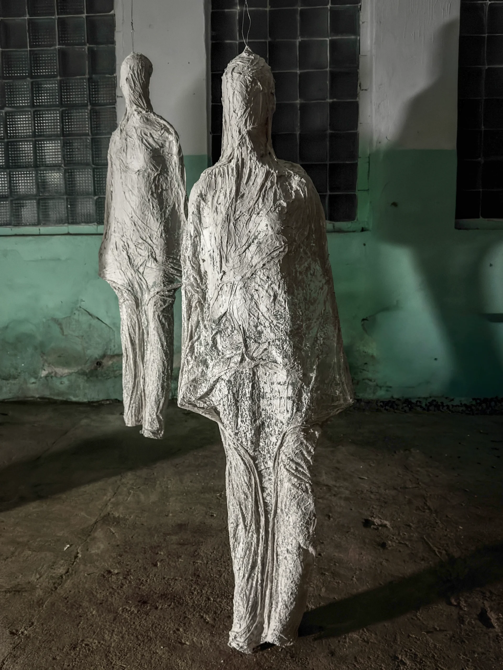 Two figures from the “Shells” installation seen close up, hanging by a glass-block window