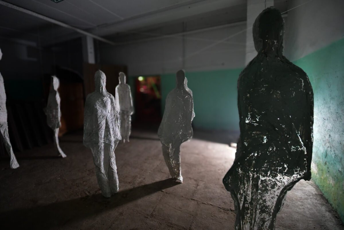 Figures from the “Shells” installation in the sawmill, with stacks of logs leaning against the wall